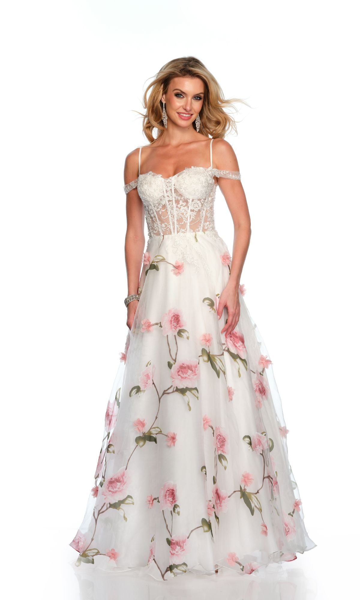 A woman wearing the Dave & Johnny Long Prom Dress 11656, a white floral gown with pink patterns, off-the-shoulder straps, a sweetheart neckline, lace bodice, and a sheer overlay stands against a plain white background.